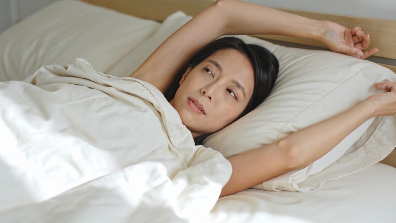 羽毛布団、枕、ヘッドボードに当たる光に促され、ベッドで伸びをして目を開ける目覚めのアジア人女性。