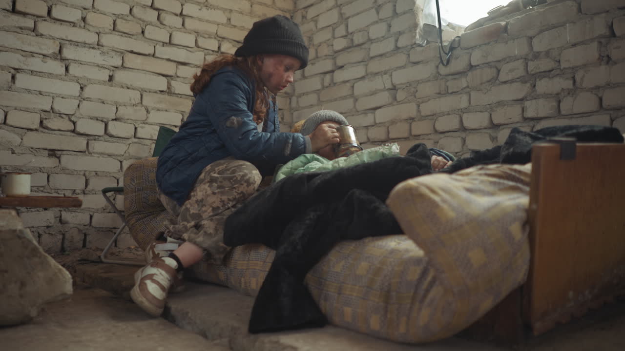 Una niña con la cara sucia, chaqueta azul y gorro ofrece un vaso de agua en un recipiente metálico a su hermana pequeña enferma, que descansa sobre un colchón desgastado dentro de un refugio frío de ladrillo, con el suelo en mal estado, provisiones esparcidas y supervivencia en condiciones difíciles.