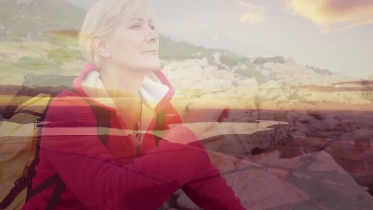 Caucasian senior woman hiking sitting on rocks relaxing, over moving clouds and sunset