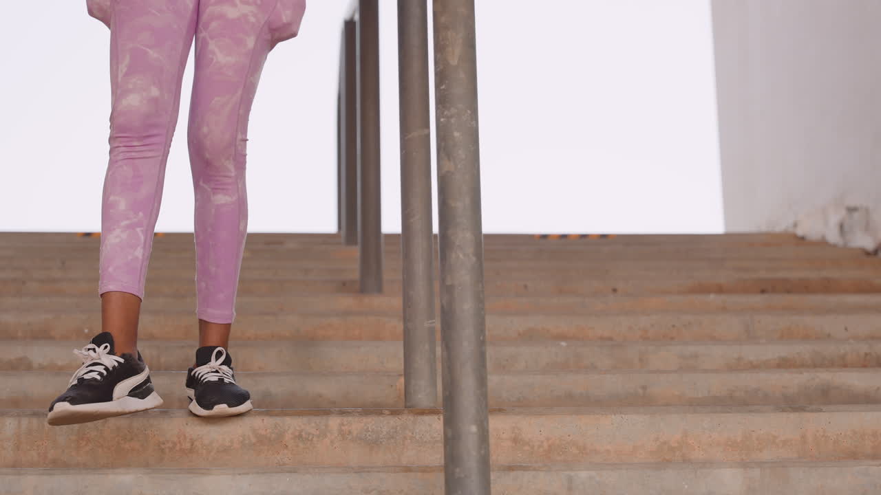 black woman stepping up stairs, closeup legs in pink leggings and sneakers, rhythmic stride on concrete steps, metal railing framing scene, urban training vibe, dance rehearsal energy, confident
