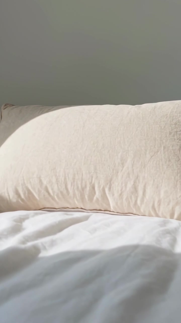 Video verticale: Luce solare che scivola su un cuscino beige su un letto bianco mentre il sole si muove, rivelando ombre.