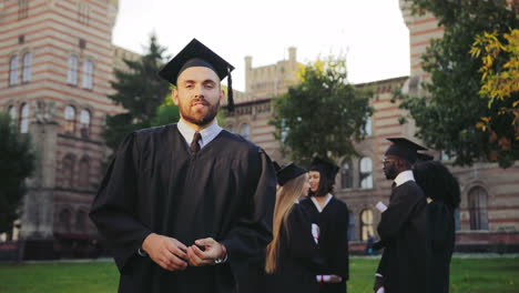 Portrait-shot-of-the-young-graduated-man-in-black-traditional-gown-and-cap-standing-in-front-of-the-camera-with-diploma-and-crossing-his-hands