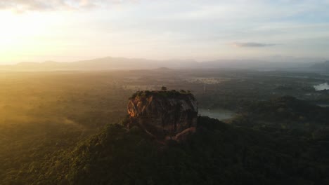 beautiful high red rock sigiriya between the green nature of sri-lanka with several blue lakes while the setting sun in the background just above mountains in the background. wide drone panning shot