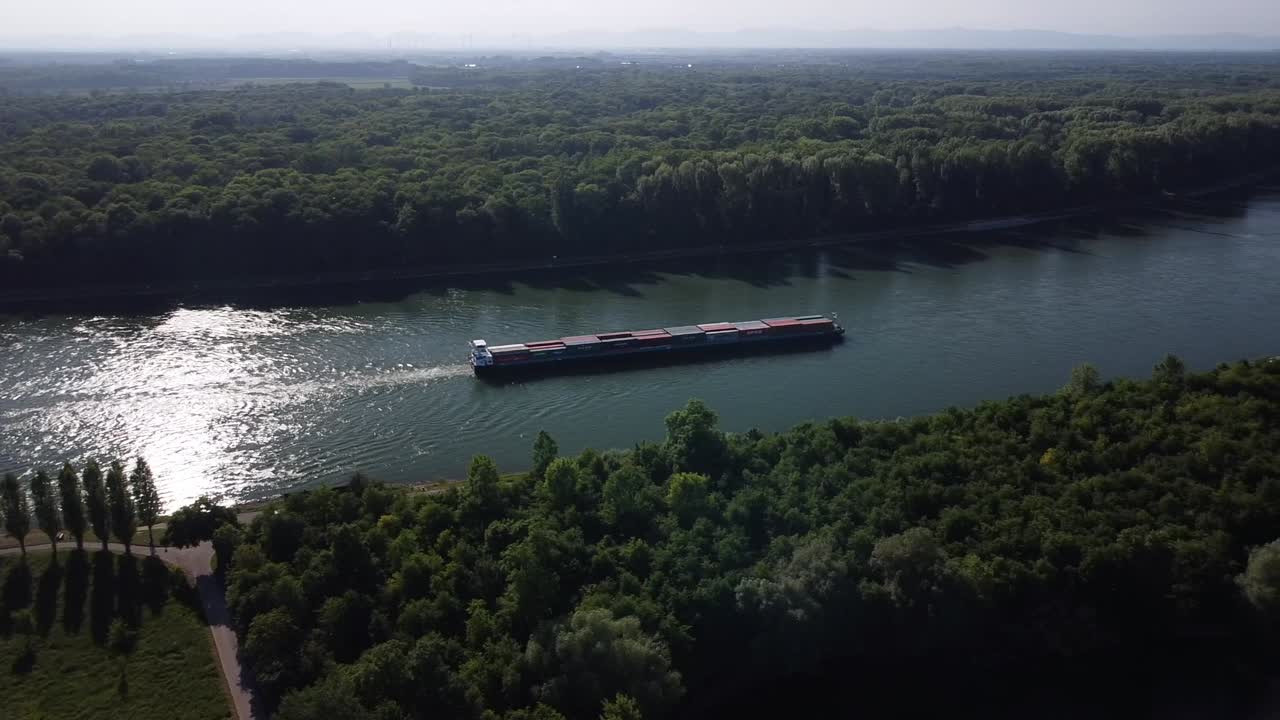 Drone camera follows a cargo ship on the Rhine River while staying in a fixed position over a beautiful forest at sunset in Europe