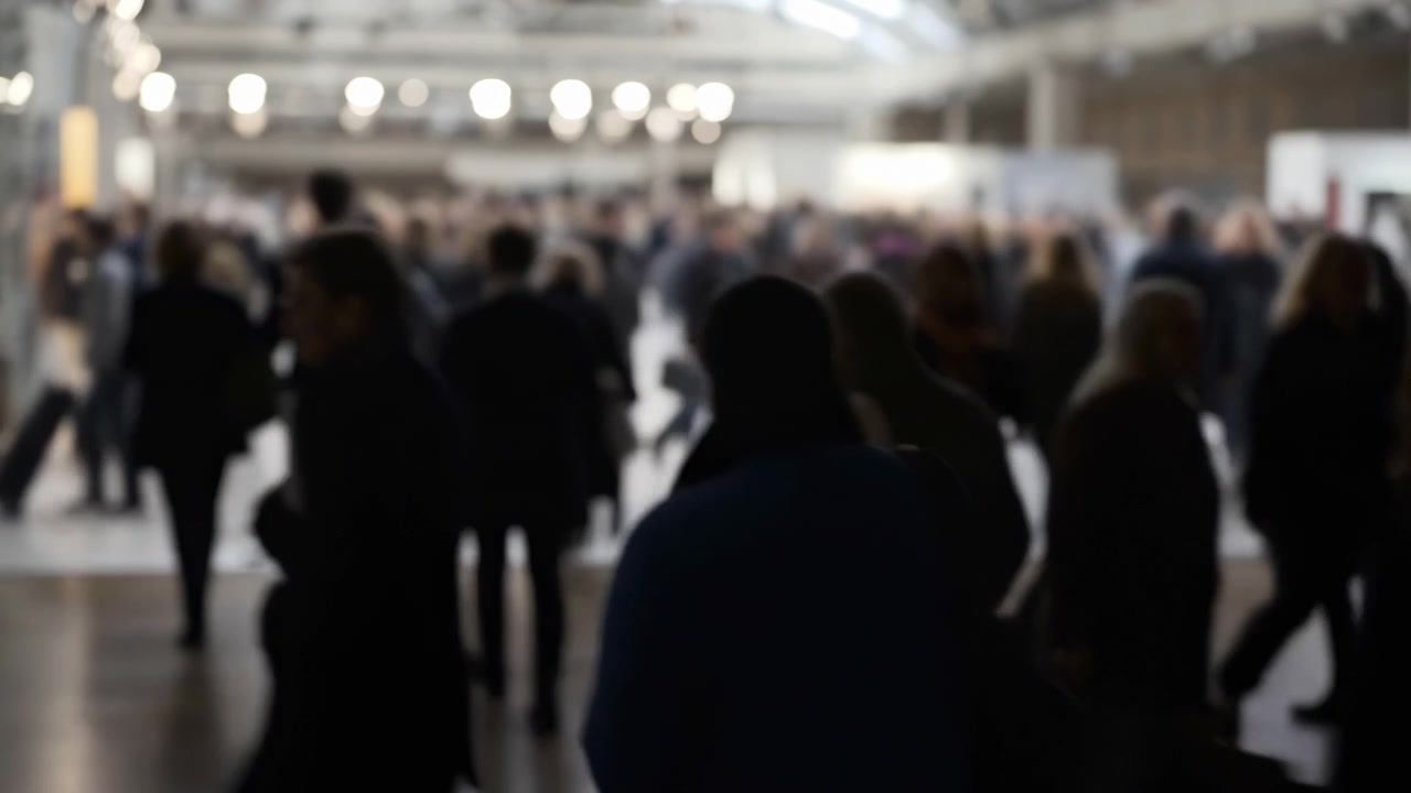 Persone in silhouette si muovono attraverso un'ampia sala espositiva interna con capriate alte e illuminazione dall'alto. Le figure in secondo piano sono scure e sfocate