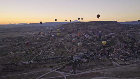 Göreme-Turkey-Aerial-V44-Flyover-Offene-Felder-Mit-Blick-Auf-Das-Dorf-çavuşin-Und-Den-Flachen-Berg-Mesa-Im-Morgengrauen-Mit-Bunten-Heißluftballons,-Die-Hoch-In-Den-Himmel-Fliegen---Aufgenommen-Mit-Mavic-3-Cine---Juli-2022