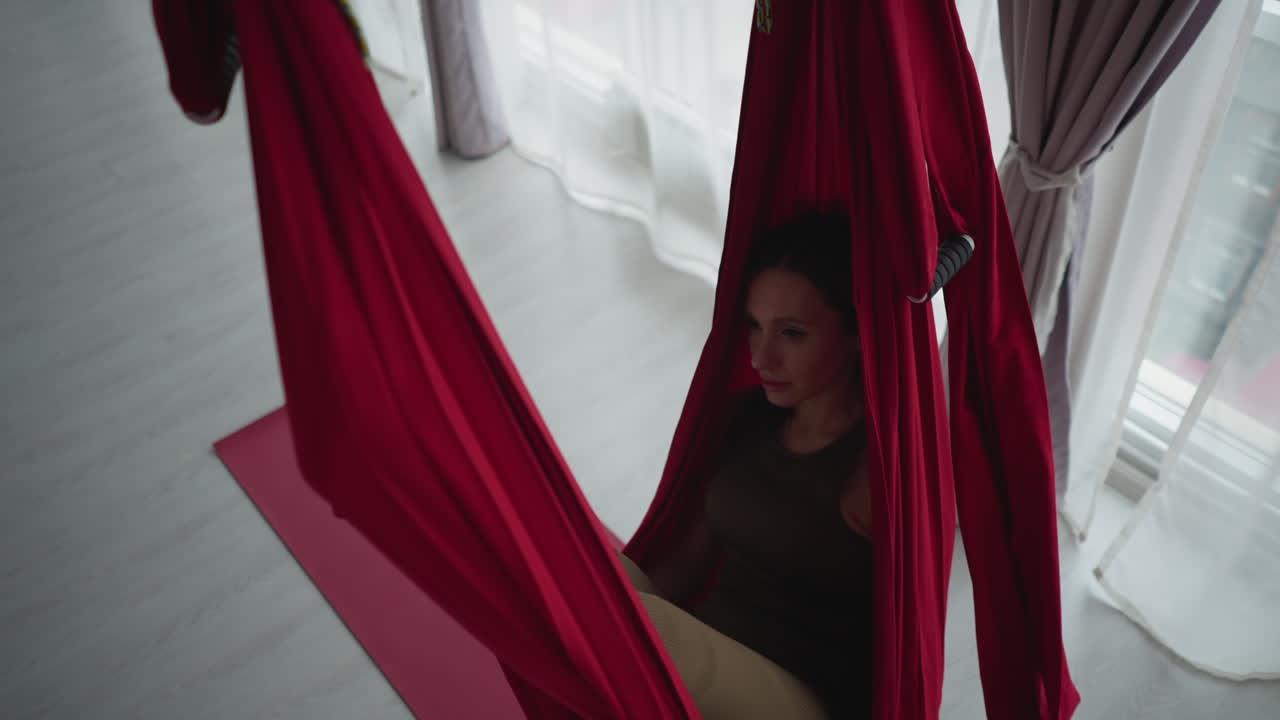 High angle view in bright glass walled yoga studio with floor to ceiling windows and sheer curtains as fitness coach lies peacefully in red silk hammock swinging gently conveying balance wellness