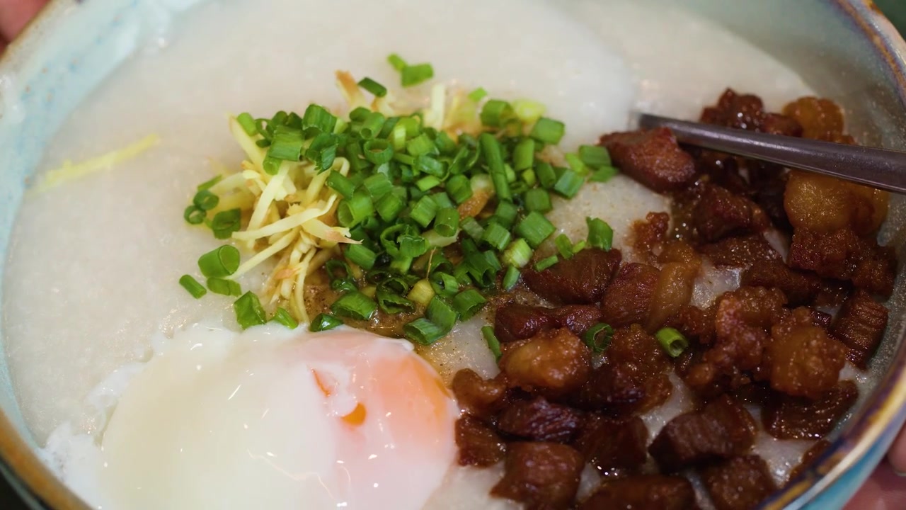 Close-up bubur nasi tradisional Asia dengan topping telur, daging babi, dan rempah-rempah dalam mangkuk