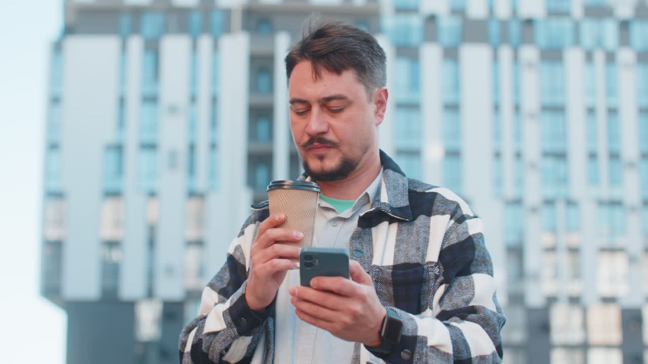 Happy man using smartphone while enjoying morning togo coffee hot drink against modern building