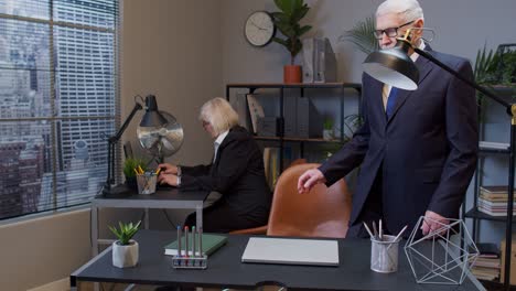 Elderly-mature-freelancer-man-walking-and-sit-at-office-workplace-with-laptop-computer-start-working