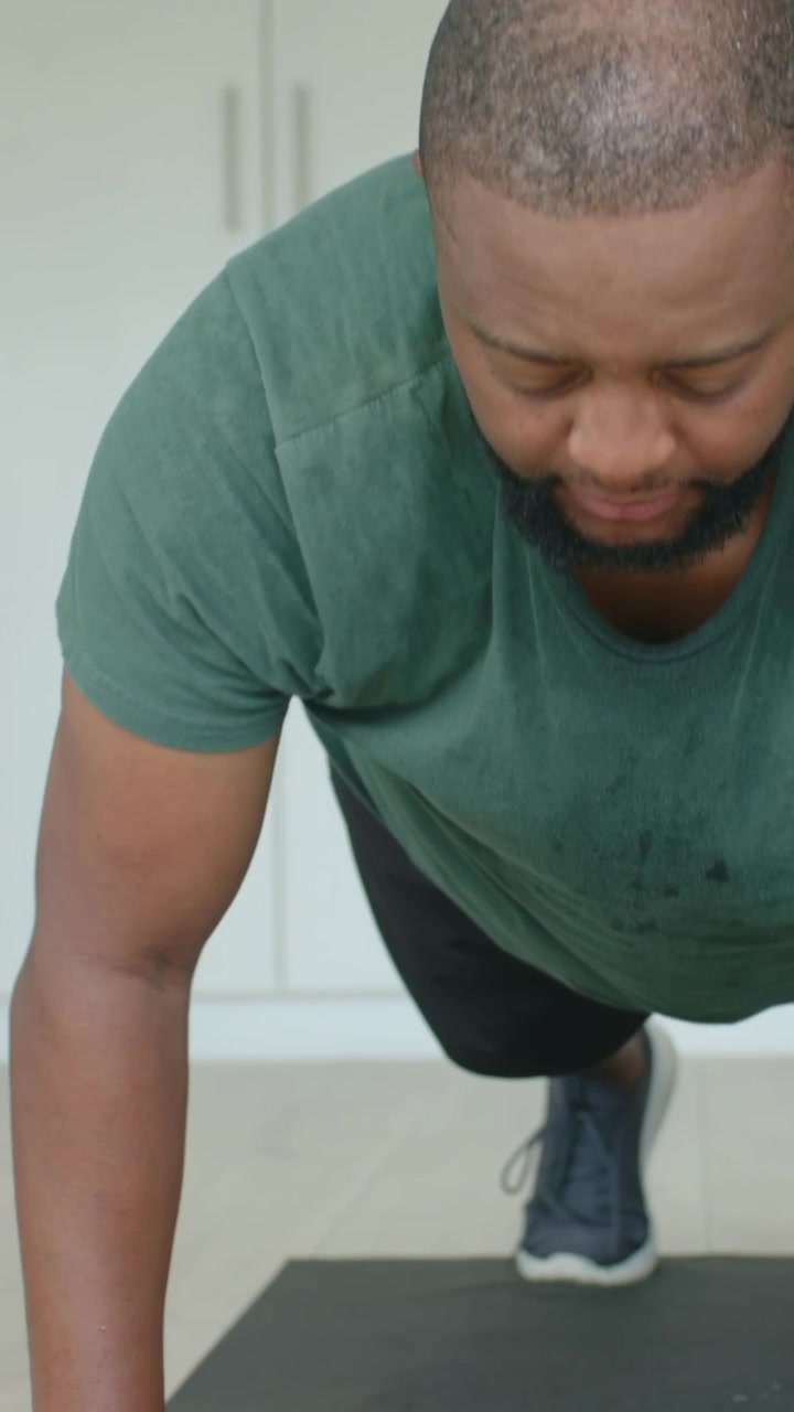 Vertical video: Performing man in green shirt starting push-up workout on black mat, with dumbbell