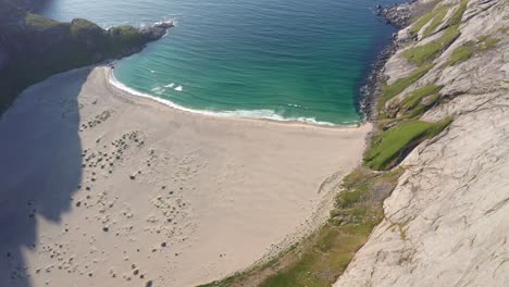 Drone-shot-of-Lofoten-steep-cliffs-and-mountains-rising-from-deep-blue-sea-with-sand-beach-bellow