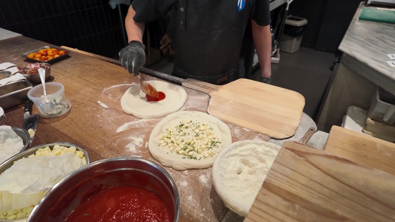 Uno chef con guanti neri applica salsa rossa su impasto di pizza crudo in una cucina.