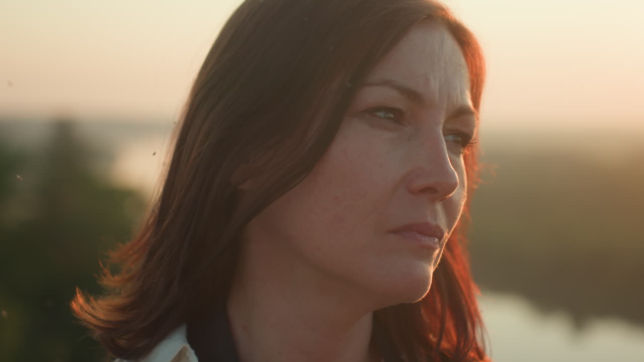 Close up of fair skin woman with distant sad gaze as insect buzzes near her in golden sunset light, soft blurred river and foliage background