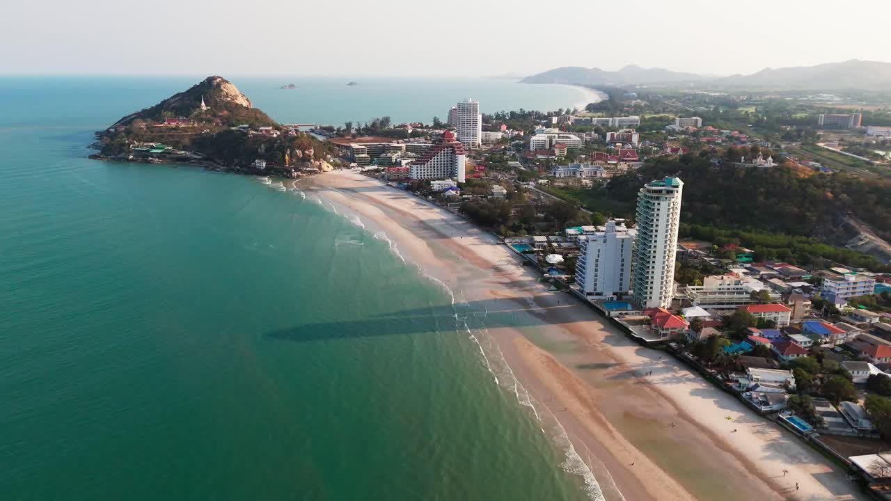 Toma aérea de dron en retroceso que muestra la costa de Hua Hin, complejos turísticos frente a la playa, olas del océano y el horizonte de la ciudad