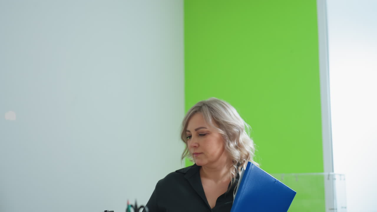 Business mogul walks into office holding coffee and blue file folder, preparing to start work, with determined expression and modern setting featuring green accent wall and bright natural lighting