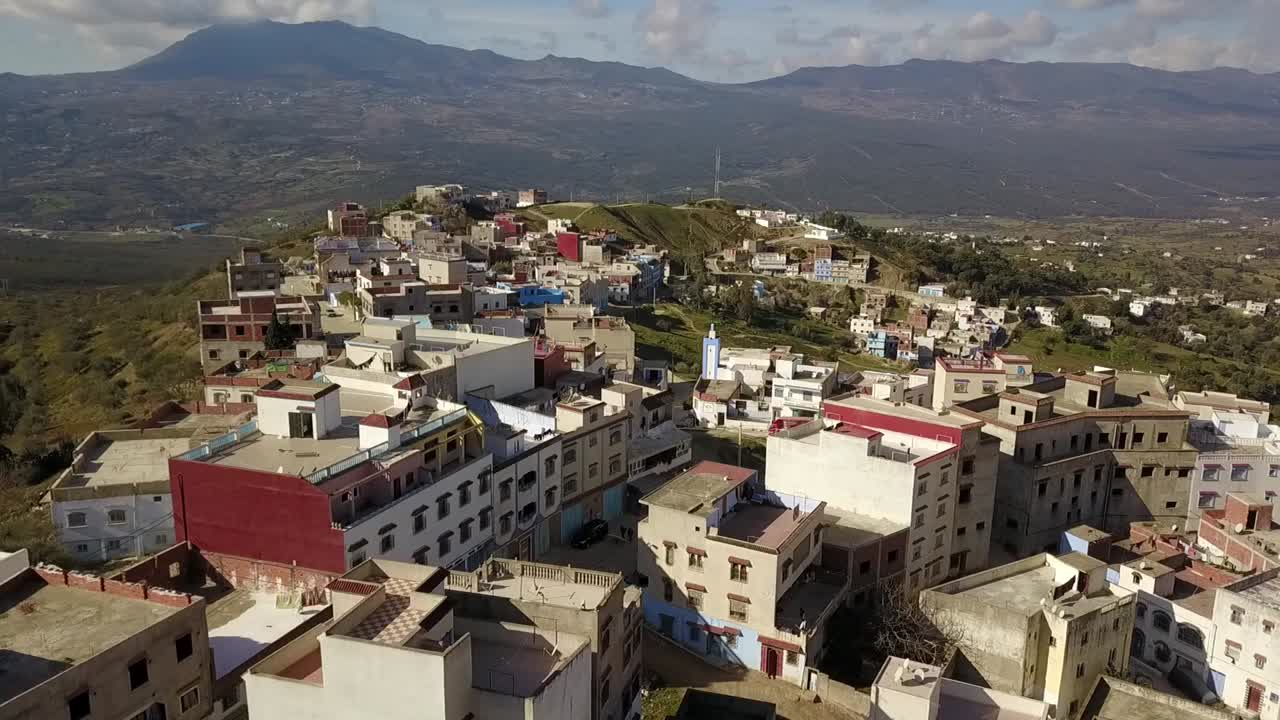 Veduta aerea di Chefchaouen, Marocco, che mostra edifici e tetti di un quartiere urbano con il paesaggio delle Montagne del Rif sullo sfondo durante il giorno.