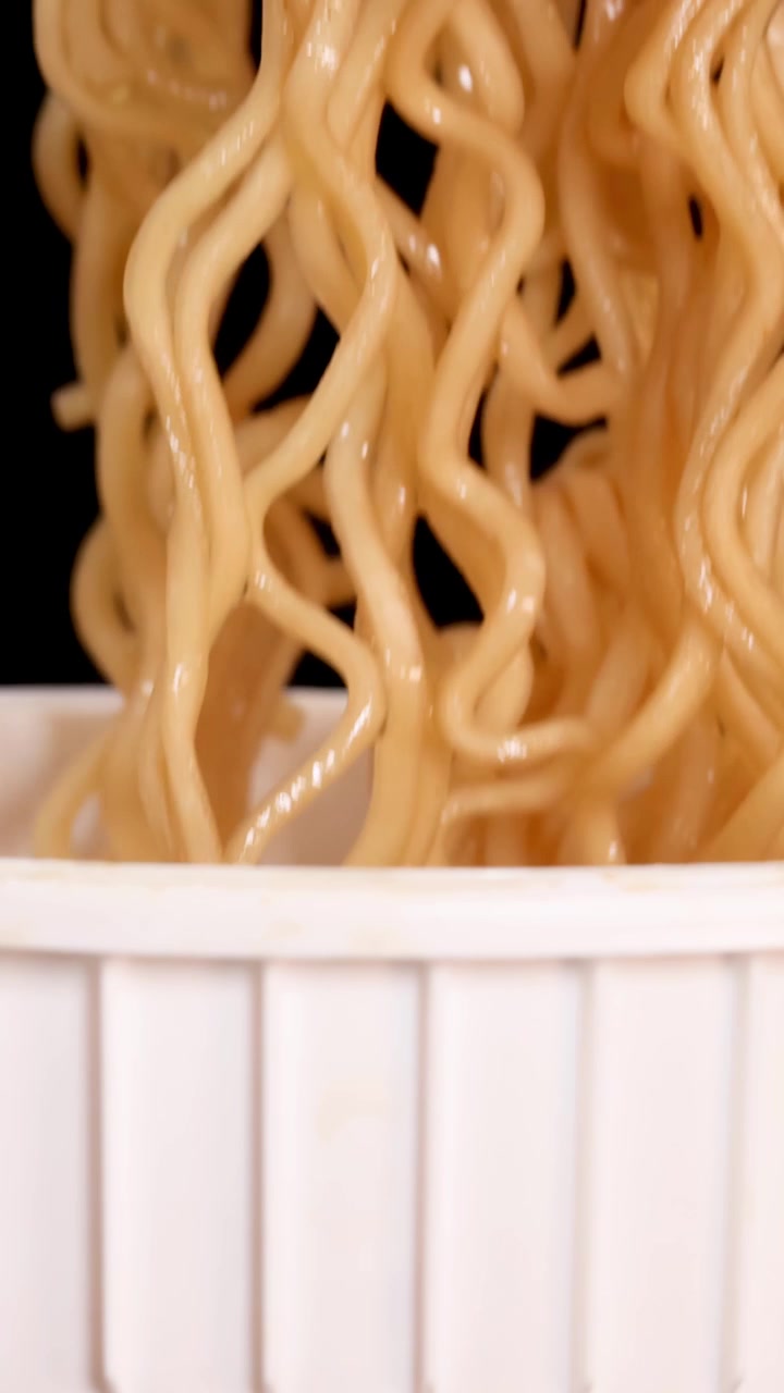 Una sequenza ravvicinata mostra spaghetti istantanei cotti sollevati da una tazza bianca con una forchetta di plastica su sfondo nero, sotto una luminosa illuminazione da studio.