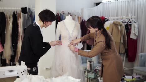 portrait of two female designers creating a wedding dress by exclusive order sewing fabrics and fixing details on a mannequin. production of wedding dresses. little business