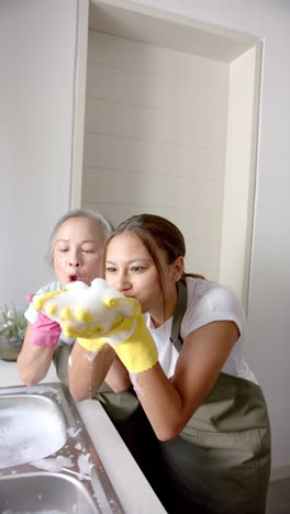 vertical video: washing dishes, woman and elderly woman having fun with soap bubbles in kitchen