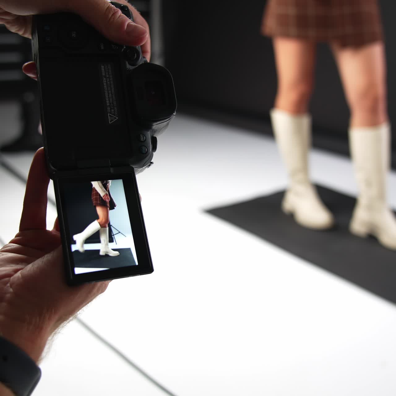 Male photographer working in studio. Camera in man's hands close up. Lady posing in white boots at backdrop in blur