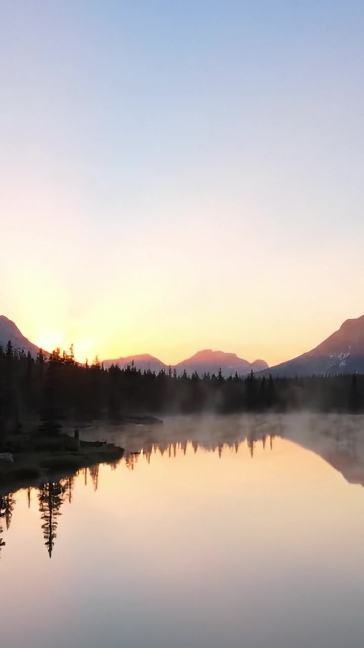 A serene dawn unfolds over a tranquil lake, capturing the soft hues of sunrise mirrored in the still water, framed by majestic mountain silhouettes and pine trees.