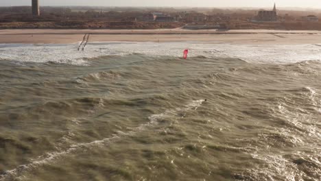 Folgeaufnahme-Eines-Kitesurfers,-Der-Auf-Einen-Strand-Mit-Einer-Touristenstadt-Im-Hintergrund-Zugeht