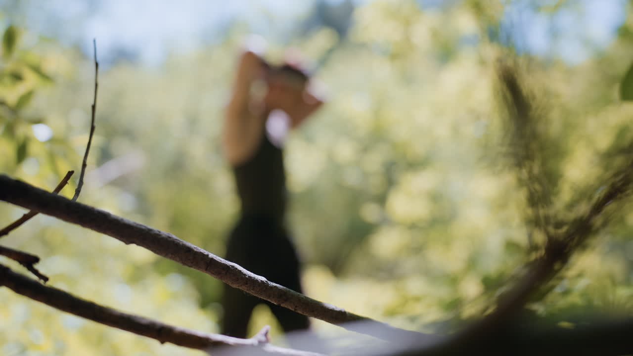 Soldier adjusting headband behind bare tree branch under dappled sunlight, emphasizing moody depth and sharp wood texture contrasting with blurred human figure wandering serene forest pathway