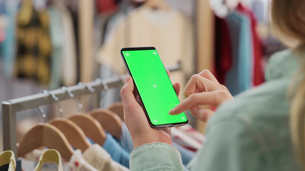 tienda de ropa: mujer usando teléfono inteligente con pantalla verde de clave de croma. colgante de ropa con elegantes artículos de marca para venta al por menor en el fondo. imágenes cercanas del dispositivo móvil.