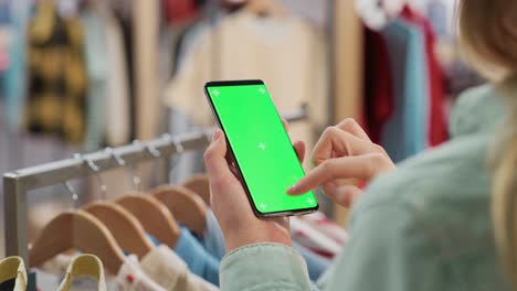 clothing store: female using smartphone with chroma key green screen display. clothes hanger with stylish branded items for retail sale in the background. close up footage of mobile device.