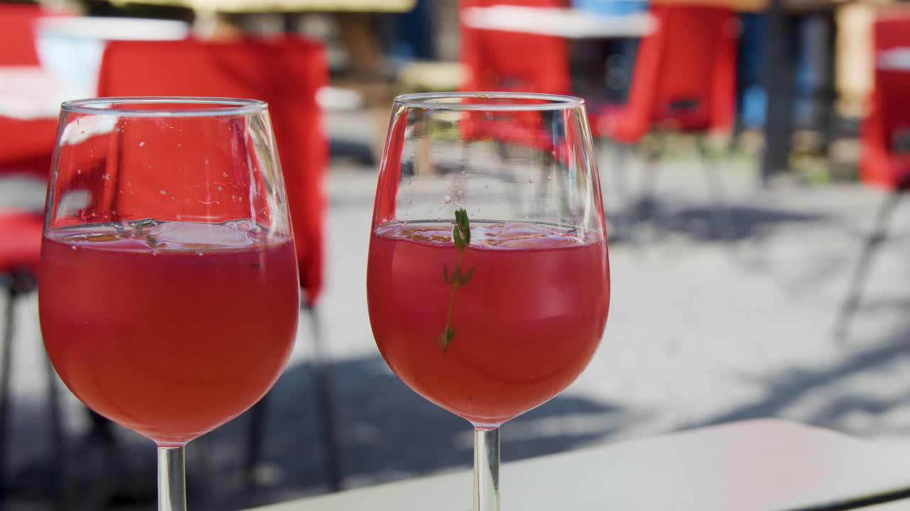 Due mani brindano con bicchieri di cocktail estivi rossi in un caffè all'aperto, sotto una luce solare brillante, con sedie rosse e un'atmosfera rilassata e celebrativa.