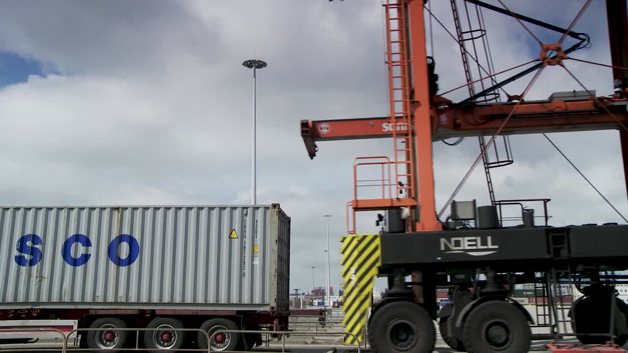 A shipping container is moved by a large crane at an industrial port under a cloudy sky