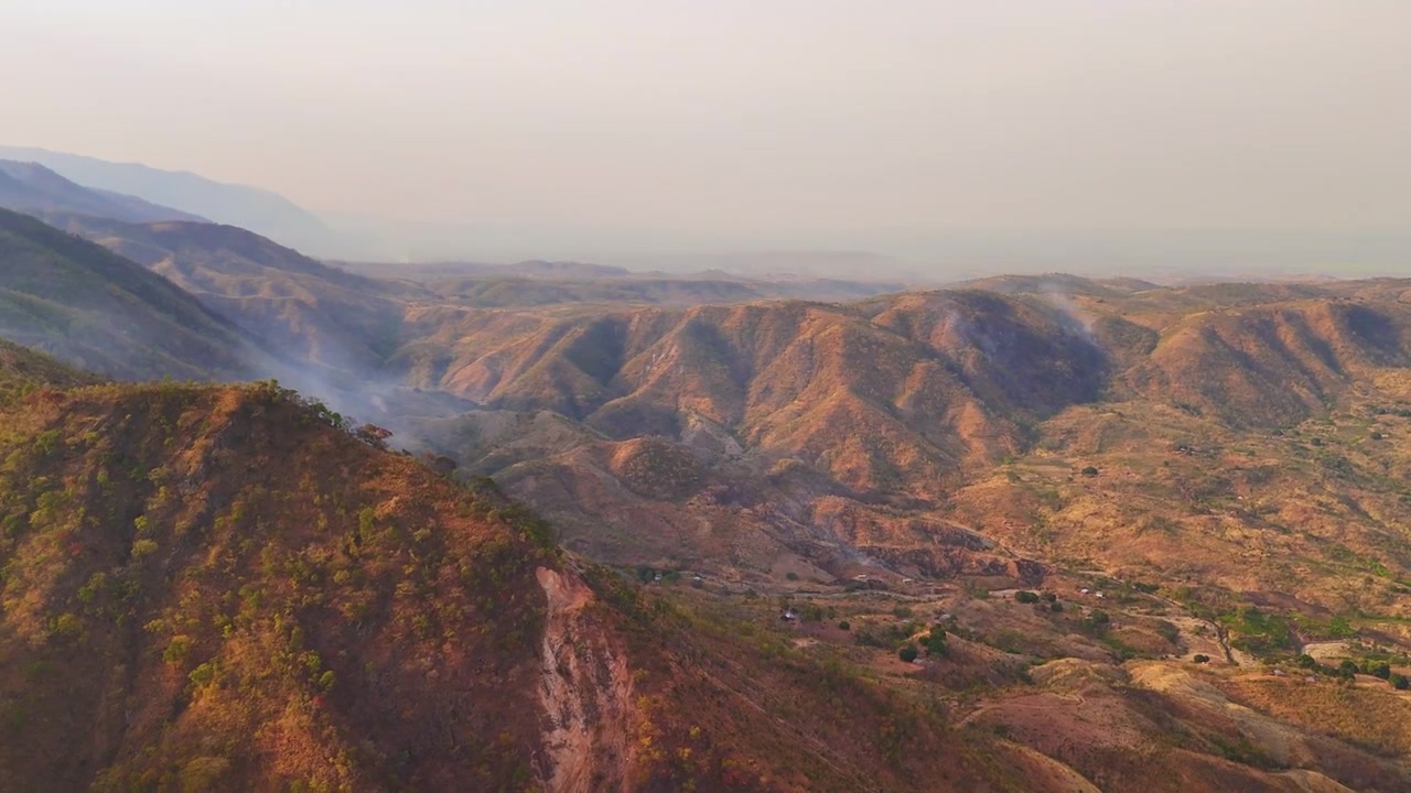Un drone sorvola gli aridi altipiani di Livingstonia alla luce del mattino, rivelando alberi sparsi, fumo in lontananza, montagne e il Lago Malawi.