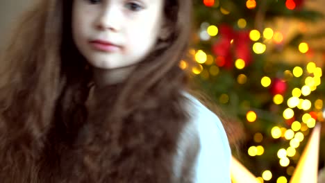 portrait of happy little girl sitting near christmas tree on new year's eve in decorated room. new year and holidays with kids concept. enjoying festive holiday celebration.