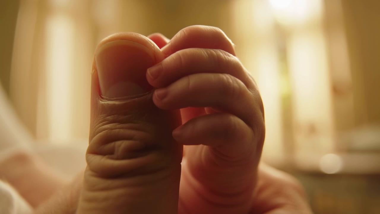 Adult thumb and fingers cradle tiny newborn toes in warm indoor light, showing delicate skin folds, soft nail detail, close personal care, intimate family bond, and calm natural texture