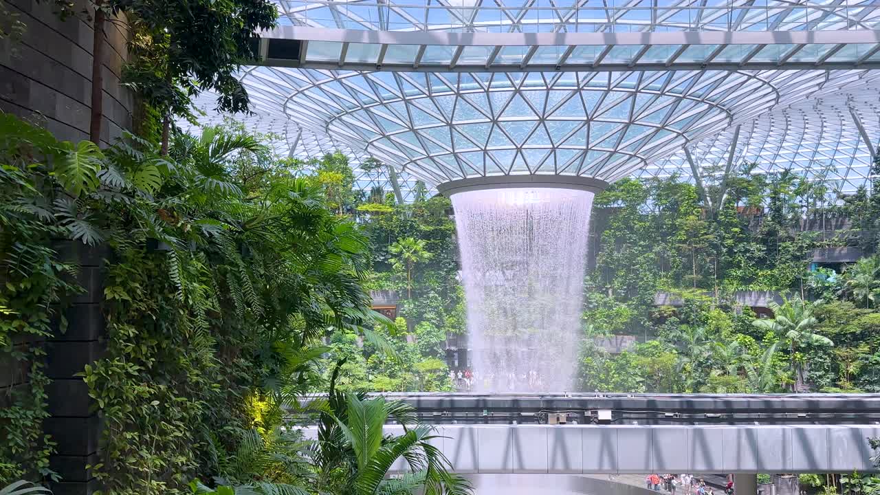 Camera slowly pans to reveal a large indoor waterfall encircled by tropical plants under a geometric glass dome, with bright natural daylight illuminating the scene