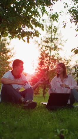dos personas sentadas al aire libre con una computadora portátil delante de ellos, brindando con rebanadas de pan en las manos, disfrutando de la naturaleza, rodeadas de árboles con la cálida luz del sol filtrándose a través