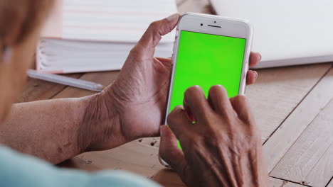 Over-shoulder-view-of-a-senior-black-woman-sitting-at-a-wooden-table-using-a-smartphone,-close-up