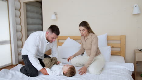 young beautiful parents sit on the bed over their little son. father raises his baby up by the feet and mom wants to kiss her child.
