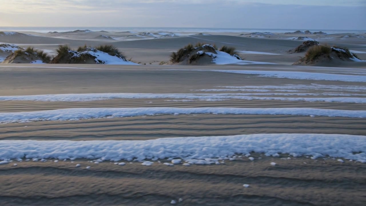Pannerende camera onthult gerimpeld zand dat verschuift bij koude kustduinen, met sneeuwstroken en graspollen