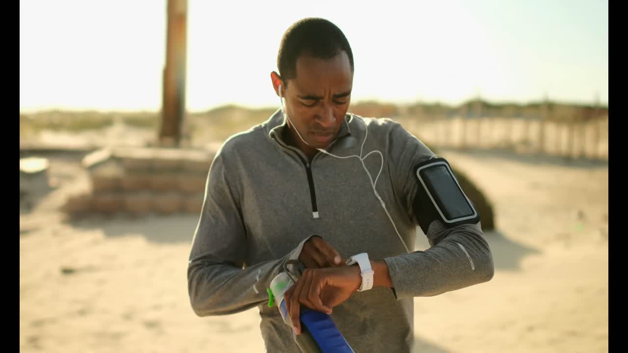 African American man wearing running arm band and using smartwatch at beach 4k