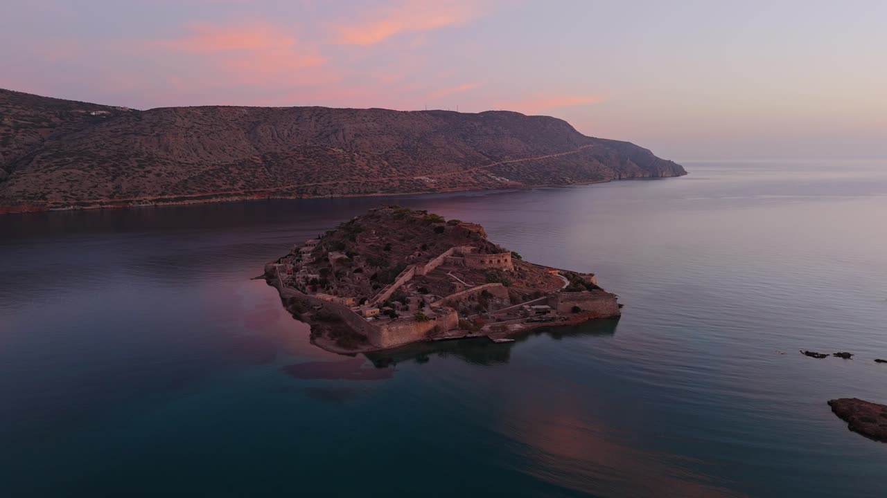 Aerial drone pullback at sunset above Spinalonga island fortress ruins below soft glowing clouds