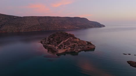 aerial drone pullback at sunset above spinalonga island fortress ruins below soft glowing clouds