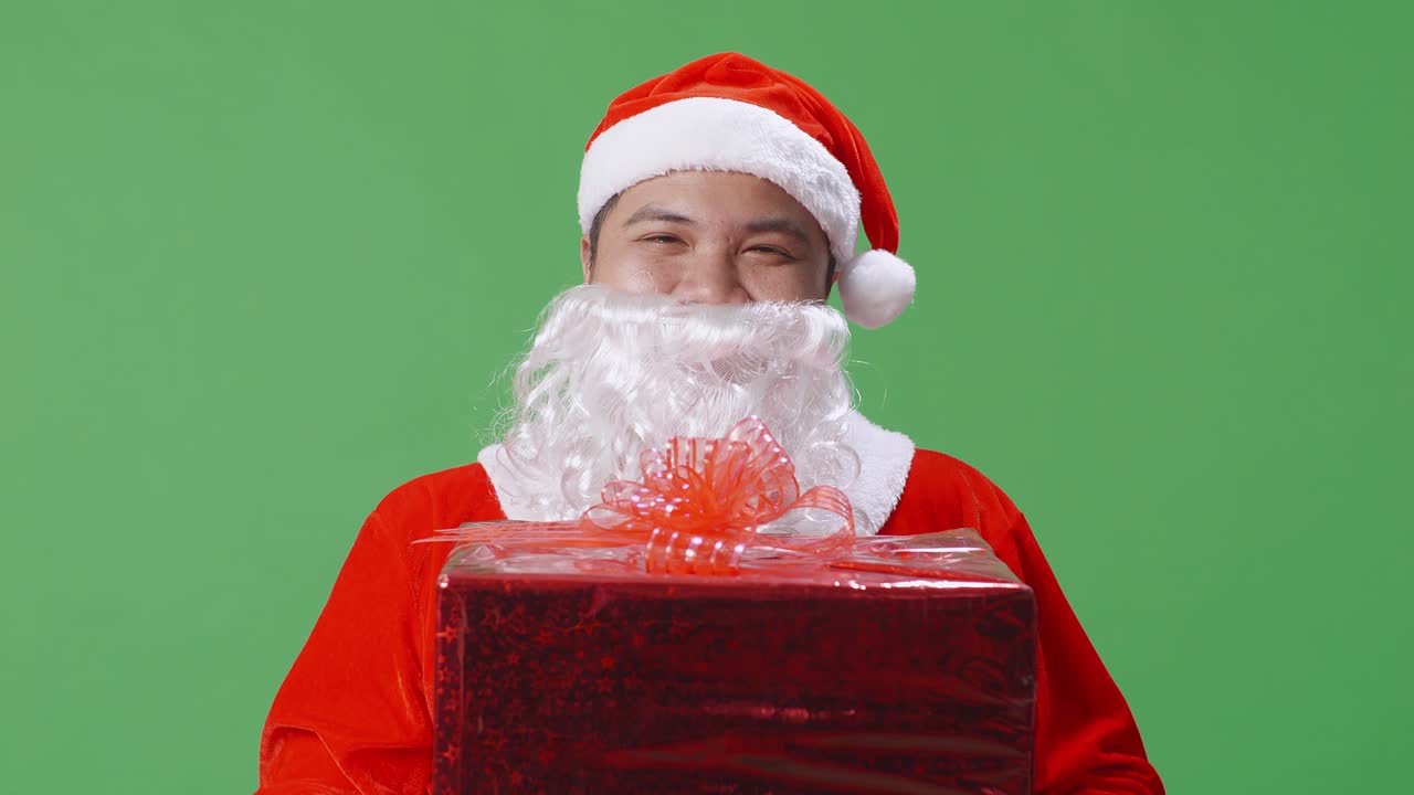 아시아 산타클로스가 스튜디오에서 녹색 스크린에 서 있는 동안 선물을 보여주고 있습니다.