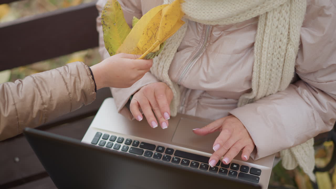 close up of woman seated on bench in autumn park typing on laptop outdoors while child reaches across keyboard to distract mother and mother gently pushes child hand away