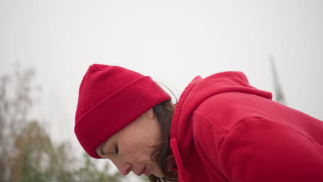 woman in red hoodie and beanie slightly bent, head dropping down slowly as she exhales, breathing out during winter in misty atmosphere with trees in background