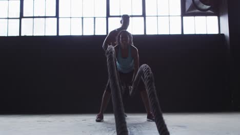 african american woman exercising battling ropes in empty urban building with man cheering her on