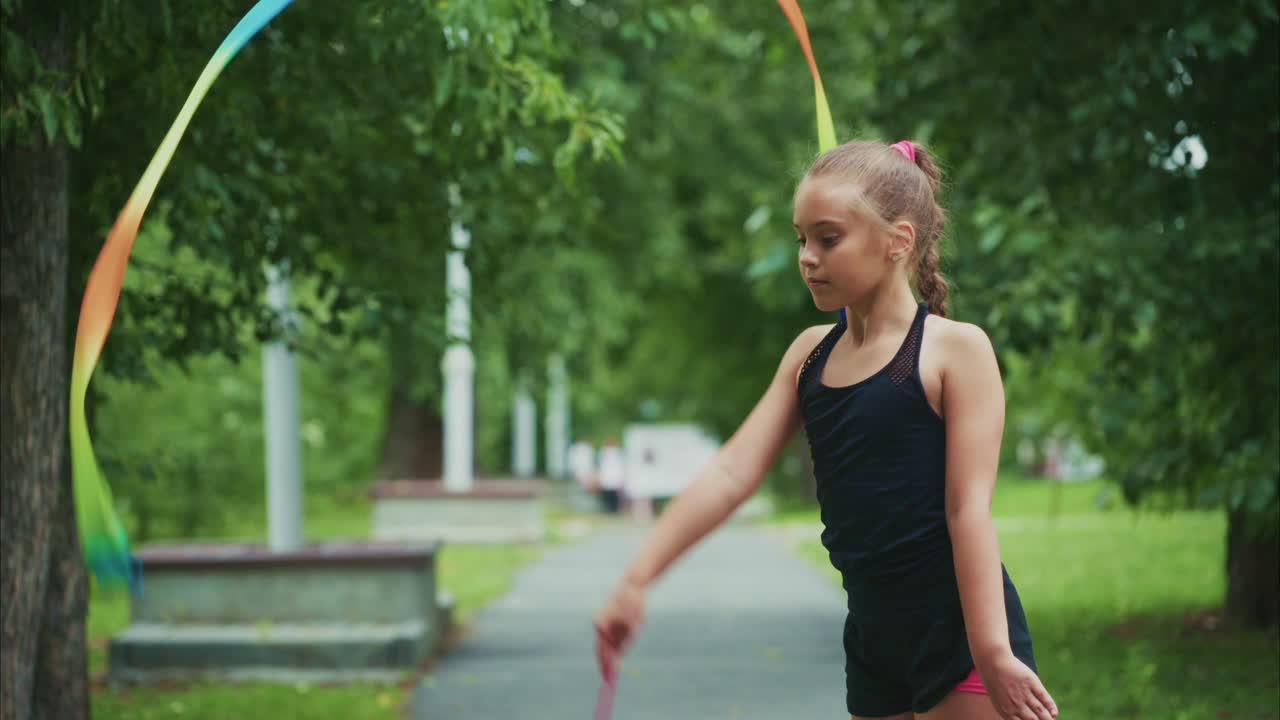 A young gymnast performs a captivating ribbon routine outdoors, showcasing grace and skill amidst a lush green park landscape, surrounded by nature