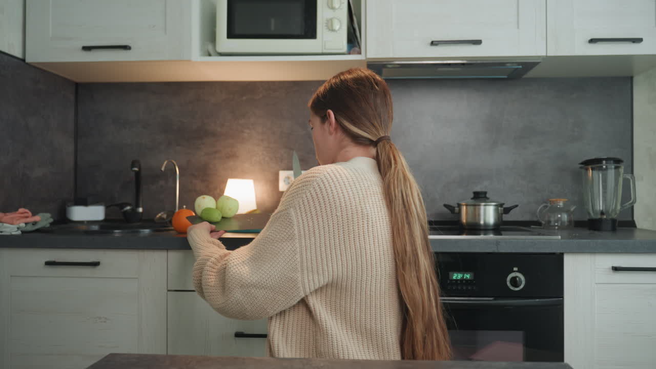 Donna bianca incinta seduta in cucina che indossa un cardigan lavorato a maglia e una bralette, mani appoggiate sul pancione visibile, luce soffusa della lampada, mele e arancia sul bancone, momento prenatale contemplativo, mattinata lenta.
