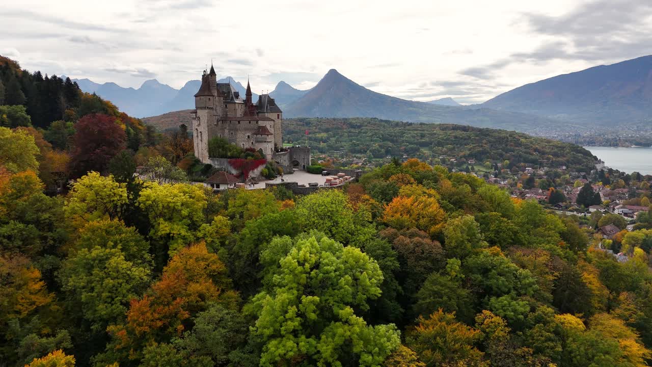 Dolly‑in aerial of Château de Menthon‑Saint‑Bernard rising above autumn woodland on eastern shore of Lake Annecy, with surrounding Bornes Massif peaks in Haute‑Savoie, Auvergne‑Rhône‑Alpes, France